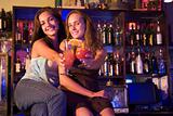 Two young women sitting on a bar counter, toasting the camera
