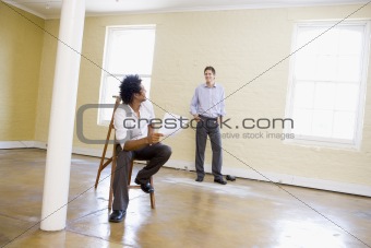 Man sitting on ladder in empty space holding paper talking to ot