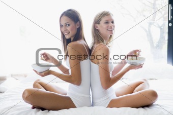 Two women sitting on bed eating cereal smiling