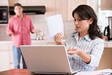 Woman in kitchen with paperwork using laptop with man in backgro