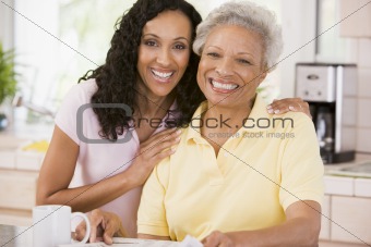 Two women in kitchen with newspaper and coffee smiling
