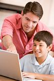 Man helping young boy in kitchen with laptop smiling