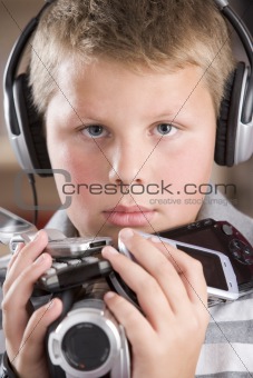 Young boy wearing headphones in bedroom holding many electronic 
