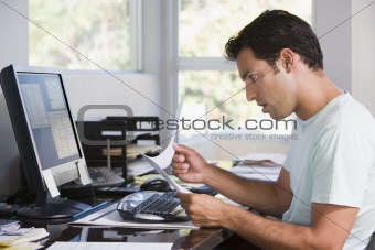 Man in home office using computer holding paperwork and looking 