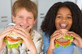 Two young children in kitchen eating cheeseburgers smiling