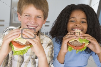 Two young children in kitchen eating cheeseburgers smiling
