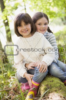 Two sisters outdoors in woods sitting on log smiling