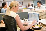 Businesswoman in cubicle using laptop and eating salad