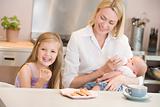 Mother feeding baby in kitchen with daughter eating cookies and 