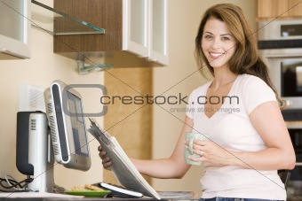 Woman in kitchen at computer with newspaper and coffee smiling