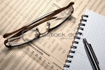 Glasses, pen and notebook laying on newspaper with financial numeric data.