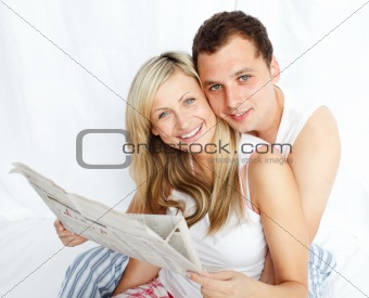 Couple reading a newspaper in bed and smiling at the camera