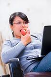 Young woman eating apple while looking at her laptop
