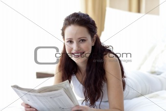 Brunette woman in bed reading a newspaper and smiling