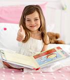 Happy little girl reading in bed with thumb up
