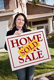 Happy Attractive Hispanic Woman Holding Sold Home For Sale Sign In Front of House.