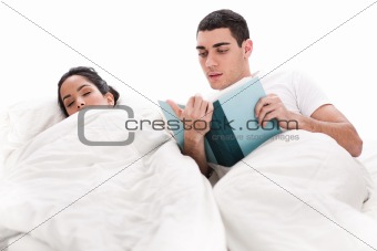 Young handsome man reading book while is wife sleeping beside him in bed
