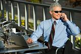 Businessman sitting at patio table ouside with laptop and talkin