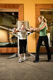 Prime adult Caucasian female helping mature adult Caucasian female use exercise equipment at gym.