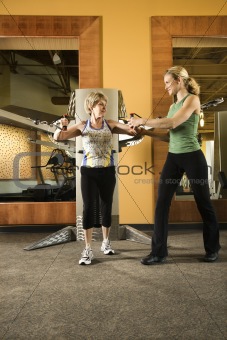 Prime adult Caucasian female helping mature adult Caucasian female use exercise equipment at gym.