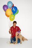 Man sitting with dog wearing party hat with balloons.