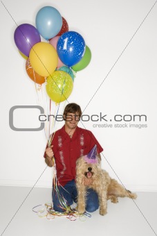 Man sitting with dog wearing party hat with balloons.