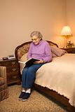 Elderly Caucasian woman reading book in bedroom.