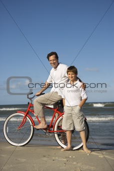 Father on bike with arm around son on beach. 