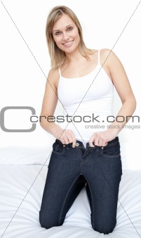 Smiling woman putting on tight jeans on a bed