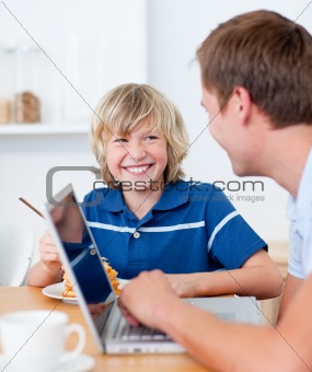 Smiling boy having breakfast while his father using a laptop