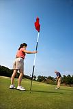 Man putting at golf course with womann holding flag.