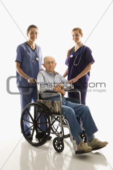 Two women wearing scrubs with elderly man in wheelchair.