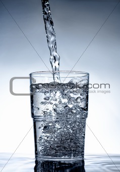Neat glass standing on water with liquid pouring into it on a beautiful background.