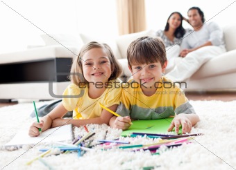 Cute children drawing lying on the floor with their parents in the background