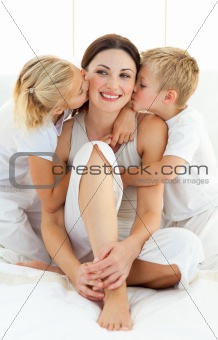 Jolly siblings kissing their mother sitting on a bed 