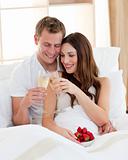 Cheerful couple drinking champagne with strawberries lying in be