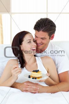 Joyful couple eating pancakes lying on their bed