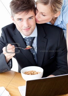 Smiling businessman eating breakfast while wife kissing his chee