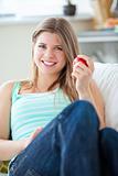 Radiant young woman holding a red apple lying on a sofa