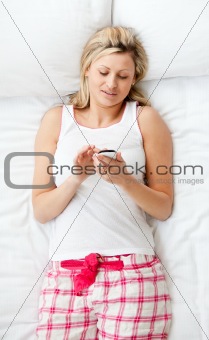 Bright woman sending a text lying on a bed at home