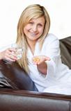 Happy woman holding pills and a glass of water smiling at the camera sitting on the sofa