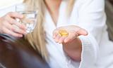 Cute woman holding pills and a glass of water smiling at the camera sitting on the sofa