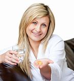 Woman holding pills and a glass of water smiling at the camera sitting on the sofa