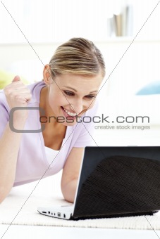 Delighted young woman looking at her laptop lying on the floor 