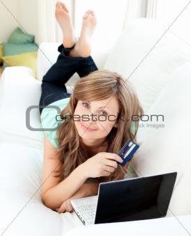 Smiling woman lying ona sofa holding a card and laptop 