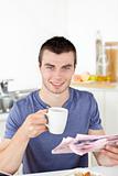 Positive young man holding a cup and a newspaper smiling at the 