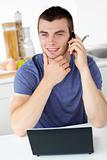 Smiling man talking on phone using his laptop in the kitchen