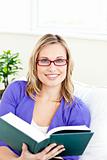Smiling young woman wearing red glasses reading a book on a sofa
