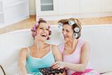 Laughing female friends with hair rollers eating chocolate at ho