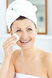 Smiling young woman with a towel putting cream on her face in th